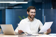 © Tetiana - A young businessman sits in the office at a desk with a laptop and looks at the documents he holds in his hands, conducting analysis and examination