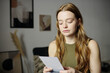 © pressmaster - Young woman with long hair is holding and reading letter in modern living room with abstract wall art and dried plant decoration, showing thoughtful expression