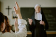 © AnnaStills - Blurred nun standing in front of classroom while students raising hands for participation. Classroom decorated with religious symbols and chalkboard in the background