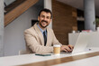 © Dusan Petkovic - Portrait of happy white collar worker sitting at office in firm and typing on laptop while smiling at the camera.