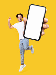 Poster - Yes, Cool Mobile Offer Ad. Joyful young casual guy showing big empty cell phone screen jumping up in the air on orange studio background, creative collage. Happy man recommend website, free copy space