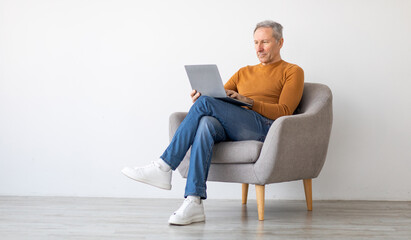 Poster - Portrait of focused mature man using laptop sitting on armchair. Confident male adult working on pc, typing on keyboard isolated on white studio background wall, full body length, free space mockup