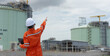 © eakgrungenerd - Professional women engineer wearing orange suit and white hard hat stands with tablet in hand at liquefied natural gas facility industrial structures petroleum.