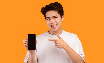 Poster - I Recommend This Website. Smiling young asian man holding smartphone with black empty screen in hand and pointing at device. Gadget with blank space for mockup, isolated over orange studio background