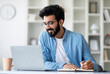 © Prostock-studio - Online Lesson. Smiling Young Indian Man Using Laptop And Taking Notes While Sitting At Desk At Home, Smiling Eastern Male Enjoying Remote Education And Distance Learning, Free Space