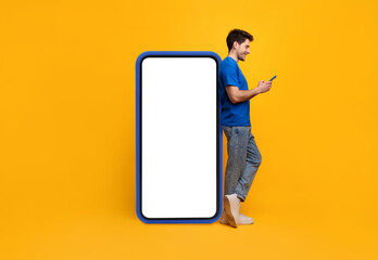 Poster - Great App. Happy young guy using cell standing leaning on big white smartphone screen with blue case on yellow studio wall, full body length. Mock up for website app, free copy space for advertisement