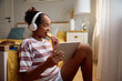 © Drazen - Happy black girl eating cookie while using digital tablet at home.