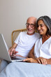 © Daniel - Vertical portrait of mature happy heterosexual couple using laptop together relaxing sitting on the bed.