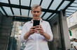 © BullRun - Serious young Caucasian freelancer using smartphone in urban area, concentrating on important message, dressed in white shirt and jeans, symbolizing digital business, finance, and career freedom