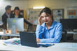 © Crystal - Young business woman suffering from headache while using laptop computer working late at night in modern office with colleagues working in background