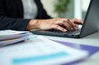 © Vera - The focus is on the movement: a businesswoman's hands typing on a digital laptop amidst a scattered stack of colorful invoices on her desk, seen in a close-up shot.