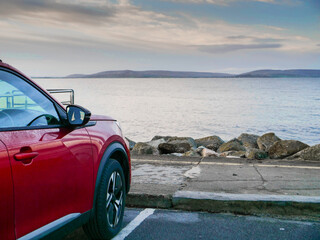 Wall Mural - Red car parked in car park with stunning view on nature scenery with ocean and mountains and cloudy sky. Silverstrand area, Galway city, Ireland. Rent motor for a tourist trip concept.