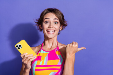 Poster - Portrait of young toothy smiling lady wear pink crop top direct finger mockup website ad for smartphone isolated on violet color background