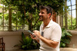 © we.bond.creations - Happy man smiling while using his smartphone in a vibrant botanical garden, enjoying nature and connecting with friends online