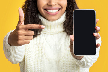 Poster - Mobile Application. Black lady in winter hat holding and pointing at empty cellphone screen over orange studio background. Happy woman showing and indicating device display for mock up
