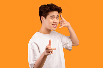 Poster - Playful asian man pointing index finger at camera, doing phone gesture saying call me back, talking with hand near ear, standing and posing isolated over orange studio background wall