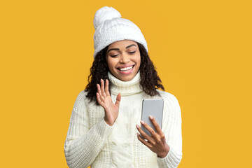 Poster - Video Call. Smiling African American Woman In Winter Hat Holding Gadget Waving Hello To Smartphone Communicating With Distant Friend Over Orange Studio Background. Modern Online Communication Concept