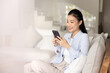 © fizkes - Young 30s Asian woman relaxing on couch, using smartphone, reading articles or online content, exploring information, engaging with communities related to hobby, enjoy new mobile application for fun