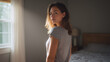 © Ilya - Young woman looking back while standing in bedroom with natural light