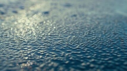 Poster - Close-up abstract of water condensation droplets on a smooth blue surface glistening in the sunlight at dawn creating a bokeh effect.