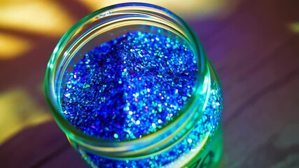 Poster - Top-down view of a clear glass jar filled with vibrant blue and purple holographic glitter on wood surface with soft lighting.