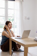 © David - Young asian woman wearing headphones and works taking notes on a Laptop while participating in a video conference on her laptop at home