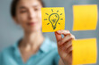© Pukan - A woman holds a yellow sticky note with a lightbulb drawing, symbolizing a new idea or inspiration, with other notes visible nearby.