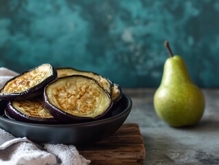 Poster - Roasted eggplant slices in a black bowl with a pear on a wooden board.