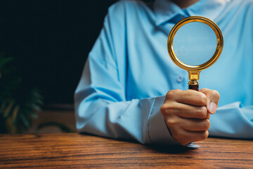 Poster - person holding golden magnifying glass on wooden table in brightly lit workspace environment