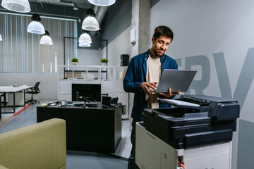 Poster - Man using a laptop while standing next to a printer in a modern office setting during daytime