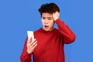 Poster - Oh No. Portrait of emotional shocked asian guy holding and using smartphone, seeing surprising news, photos or message. Surprised man grabbing head isolated over blue studio background wall