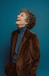 © Centric  - Elderly woman with curly hair laughing joyfully, wearing a brown suit and blue turtleneck, standing against a vibrant blue background