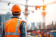 © AungThurein - A construction worker in safety vest and helmet, at building site, cranes and structure behind