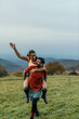© La Famiglia - Happy couple piggybacking in a meadow with mountain view: celebrating successful hike