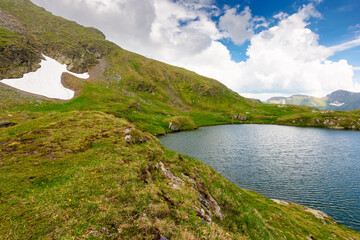 Wall Mural - alpine lake in fagaras mountains. grassy slopes with stones and snow under cloudy sky in summer. rip