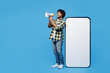 © ViDi Studio - Full body young happy Indian man he wear yellow checkered shirt casual clothes big huge blank screen mobile cell phone smartphone with area scream in megaphone isolated on plain blue cyan background.
