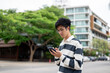 © bongkarn - Asian man wearing glasses is looking and typing at his smartphone while standing in the parking lot.