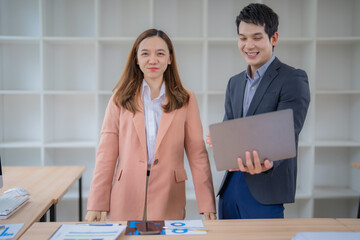Poster - Two Asian businesspeople discussing work while using a laptop in a modern office, surrounded by charts and graphs on the table, highlighting collaboration and strategy for success