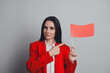 © deagreez - Successful businesswoman holding a red flag on a grey background showing a sign of alert, dressed in a red suit