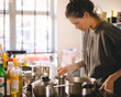 © zvkate - Woman standing by the stove in the kitchen, cooking and smelling the nice aromas from her meal in a pot. Housewife cooking lunch or breakfast.