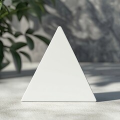 Aufkleber - A pristine white triangular prism stands on a light gray surface, subtly shadowed, with blurred foliage and a textured backdrop