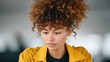 © Kamonwan - A young woman with curly hair is engaged in deep thought. She is wearing a yellow jacket and looks focused, capturing a moment of introspection in a modern workspace.