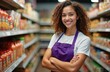 © Vadym - Smiling woman in apron stands at grocery store aisle. Fresh food on shelves behind. Smiling staff member poses looking at camera. Supermarket shopping concept.