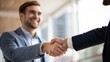 © Komdo - Two professionals in suits shake hands, smiling, in a bright office setting, symbolizing agreement or partnership.