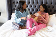 © Wavebreak Media - Sitting Diverse female friends chatting on white bed against tufted headboard, with ceramic mugs