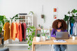 © Studio Marmellata - A woman with curly hair works on her laptop. She is managing her clothing business from her bright workspace, surrounded by colorful garments and plants.