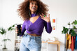 © Studio Marmellata - A joyful woman with curly hair is dancing and listening to music through wired headphones connected to her phone, expressing happiness and freedom in her home.