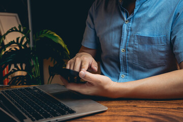 Poster - Close-up of person in blue shirt using smartphone while sitting at desk with laptop and plant in background