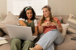 © Wavebreak Media - Smiling Diverse female friends working on silver laptop on couch in living room, with notebook