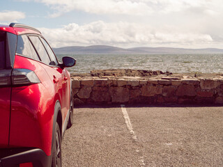 Wall Mural - A red car is parked in a parking lot next to a wall. The car is facing the ocean, and the sky is cloudy. Car park with stunning view on Galway bay and Burren mountains. Travel and tourism.